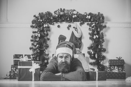 christmas man with beard on sad face in red santa claus hat near present box at new year decoration fireplace, winter holiday celebration, boxing dayの写真素材