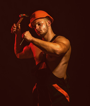 A site is also under construction. Construction worker hammer a nail. Man work with hammer. Hard worker use muscular strength. Muscular man builder at work under constructionの写真素材