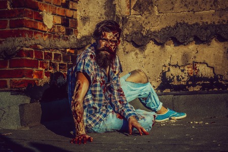 Bearded zombie man with beard creepy vampire or bloody war soldier with wounds and red blood sits on asphalt roadの写真素材