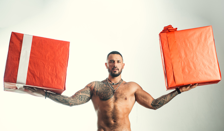 Providing fast gift delivery. Brutal latino man holding gift boxes in strong hands. Bearded hispanic man with wrapped gift boxes. Muscular shirtless guy holiday gifts in red wrapの写真素材