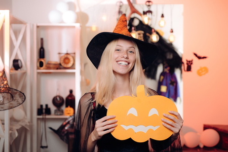 Halloween dresses and witch costumes design. Pretty young blonde woman dressed as a fairy with pumpkin. Charming halloween witch with broom over vintage background.の写真素材