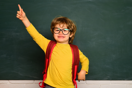 Elementary school and education. Cute boy with happy face expression near desk with school supplies. Funny little boy pointing up on blackboard.の写真素材