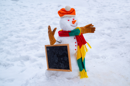 Snowman is standing in winter hat and scarf with red nose. Snowman in a scarf and hat. Greeting snowman. Cute snowmen standing in winter Christmas landscape.の写真素材