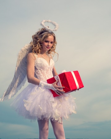 Lovely girl Cupid angel with gift with bow and arrow is ready to find your love. Portrait of little curly blond Angel girl. Cute teen Cupid angel with gift on the cloud - sky background.の写真素材