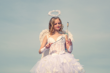 Lovely and cute youth. Girl angel with halo in white angel dress. Valentines day cupid. Cupid in valentine day. Little goddess with white wings alone on blue sky background. Arrow of love.の写真素材