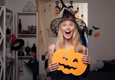 Attractive model girl in Halloween costume. Halloween Witch with pumpkin. Beautiful young woman in black witch costume with hat and broom standing over orange background.の写真素材