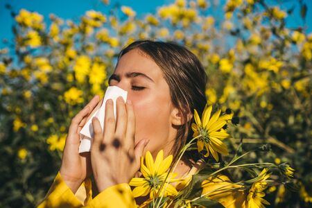 Pollen allergy, girl sneezing in a field of flowers. Jerusalem artichoke flowering. Napkin for running nose. Woman wearing yellow jacket.の写真素材