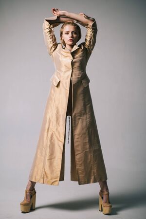 Full-length studio fashion portrait of young beautiful model wearing long white trench coat, golden heels, posing on grey background.の写真素材