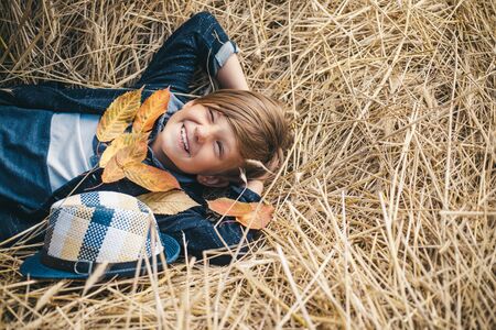 Joyful guy with a hat on his head in the hay. The boy advertises childrens clothes for the autumn. Happy childhood. Cute boy on an autumn holiday in the village.の写真素材