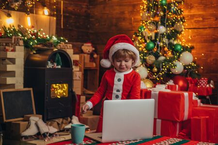 Little genius. Santa little helper. Little boy santa hat and costume having fun. Boy child with laptop near christmas tree. Buy christmas gifts online. Christmas shopping concept. Gifts serviceの写真素材