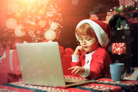 Christmas shopping concept. Gifts service. Santa little helper. Smart toddler surfing internet. Little boy santa hat and costume. Boy child with laptop near christmas tree. Buy christmas gifts onlineの写真素材