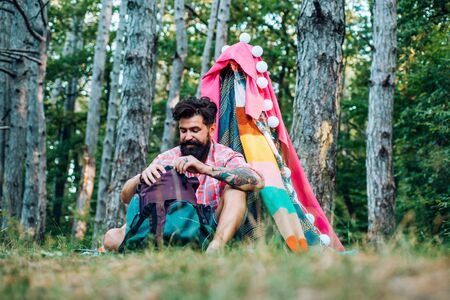 Cozy play tent for man in forest park. Hiking and outdoor recreation concept with flat camping travel. Handsome bearded man having fun in adventure Park.の写真素材