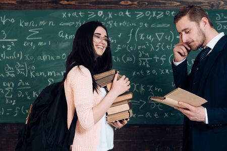 Happy woman student in glasses with book stack and backpack smile with teacher man on chalkboard in classroom. More knowledge. Bookworm. I love reading. Literature interests meの写真素材