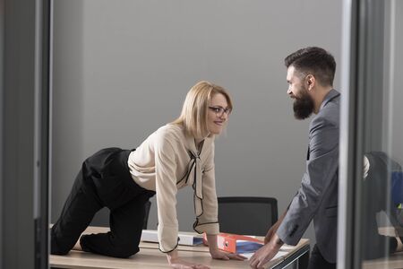 Office romance concept. Sexy secretary seduce boss in office. Businesswoman on desktop look at bearded businessman. Man under woman domination, matriarchy. Sexual flirt at workの写真素材