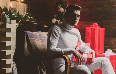 Handsome man with christmas gift. Portrait of a elegant mysterious stylish man in white clothing sitting in a chair, holding Christmas gift. Creative Marketing Ideas for 2020. Holiday promotions.の写真素材