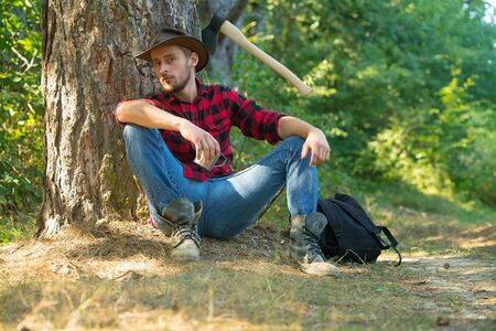 Lumberjack worker man sitting in the forest. Lumberjack with axe on forest background. Man doing mans job. Lumberjack worker standing in the forest with axe. Resting after hard work.の写真素材