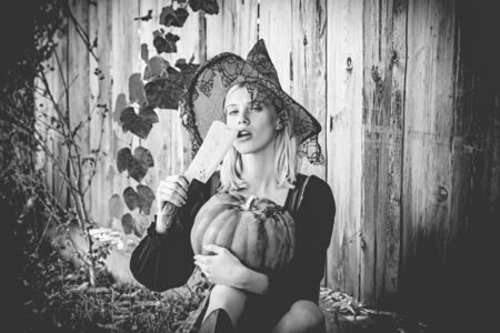 Halloween woman. Attractive witch holding pumpkin and knife for a trick or treat. Woman dressing in Halloween costume with orange witchs hat for a themed party. October festival. Wood background.の写真素材