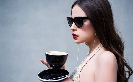 Morning coffee. Beautiful fashion model posing in glasses. Beauty trends. Female fashion model posing with cup. Young woman posing. Portrait of serious woman in the grey isolated.の写真素材