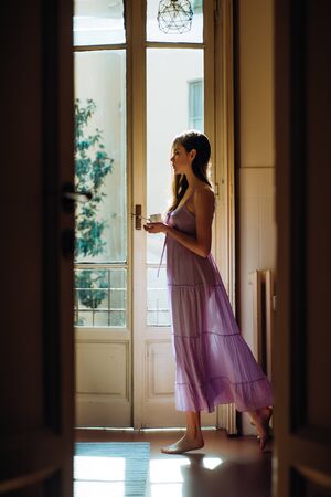 She holds a cup she happy dances. Girl is dressed in nightwear. Balcony with house view. Portrait beautiful girl with long hair. Sunrise at morning.の写真素材