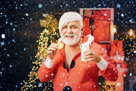 Santa in home. Santa picking cookie and glass of milk at home. Christmas Beard style. Portrait of Santa Claus Drinking milk from glass and holding cookies.の写真素材