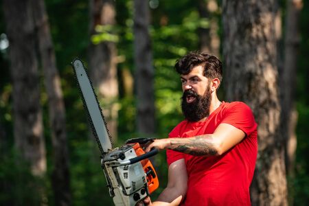 Lumberjack on serious face carries chainsaw. Woodcutter with chainsaw on sawmill. A handsome young man with a beard carries a tree. Harvest of timber.の写真素材