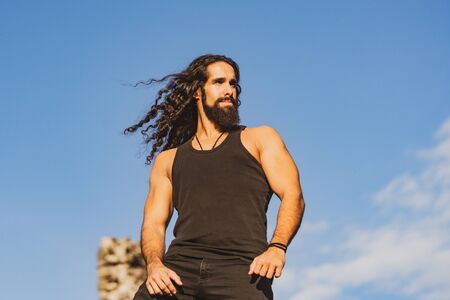 Handsome bearded man with long dark hair. Fashionable man with long wavy hair and strong healthy body. Mens style and wellness concept. Strong muscular handsome man at blue summer sky background.の写真素材
