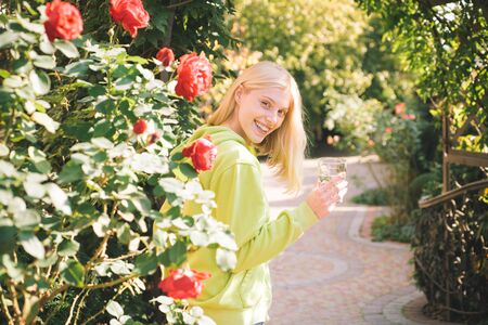 Beautiful sensual woman standing behind hot red roses and holding glass of fresh water. Active lifestyle at autumnal park. Fall leafs background. Sunny warm autumn day. Autumn concept.の写真素材