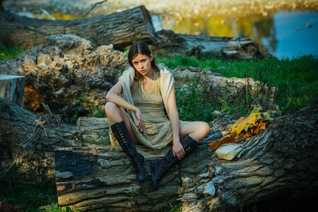 Serious woman on trunk wood. Adult girl relaxed sitting on wood in an outdoor park. Portrait of a beautiful charming young attractive woman in a dress. Girl enjoying nature.の写真素材