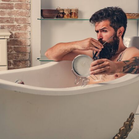 Grooming beard. Bearded man with beard with mirror in hand. Morning hygiene. Skincare in home bathroom.の写真素材