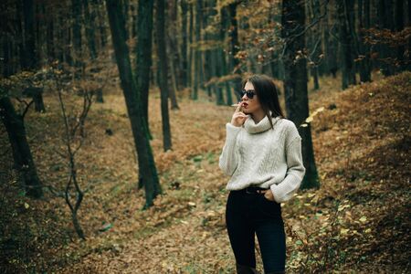 Woman walking at autumn nature in autumnal stylish outfit. Stylish femme fatale wearing fashionable clothes and smokes a cigarette at fall forest background.の写真素材