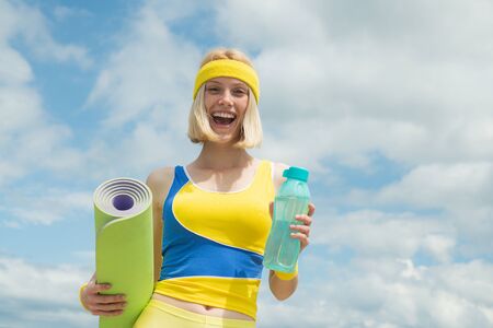 Water and hydration concept. Sport activity outdoor. Blonde cheerful smiling woman holding sport yoga mat and bottle with water. Water is vital necessity during sport exercises.の写真素材