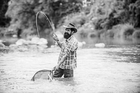 Bearded fisher in water. Young man fishing. Successful fly fishing. Fisher masculine hobby. Man with fishing rod and net. Master baiter. Fishing. Real happiness. Active sunny day.の写真素材