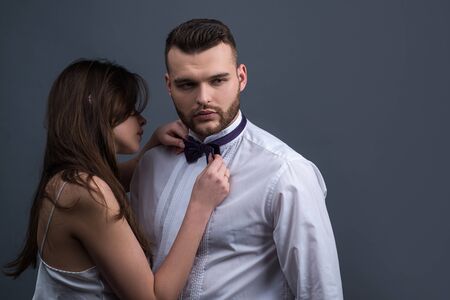 Portrait of elegant couple posing together on grey studio background. Elegant modern male accessory. Women holding bowtie ready to dress it on man in white shirt. Couple getting ready for event.の写真素材