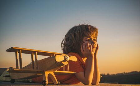 Child dreams and imagination concept. Dreaming child playing with toy wooden airplane against summer...