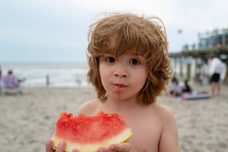 Funny portrait of an incredibly beautiful Red-haired little boy eating watermelon. Healthy fruit snack, adorable toddler child with curly hair on summer beach.の写真素材