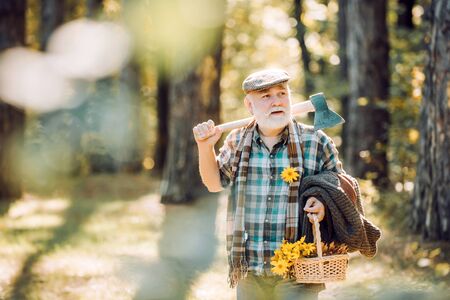 Bearded man relax in forest. Smiling old forester with axe and flowers. Happy man with beard and mustache hold axe. Elderly male walk in forest. Bearded man in hat on a background of trees.の写真素材