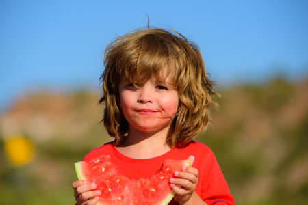 Boy child eating watermelon with funny face, green nature background. Close-up photo of kids eating sweet red melon.の写真素材