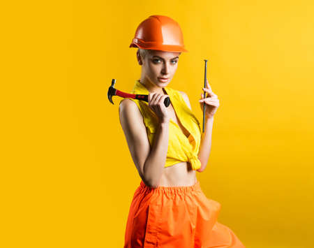 Sexy woman in helmet to use hammer. Young woman hammering nail at workshop on yellow background isolated. Female model working on building. Everything can be fixed concept.の写真素材