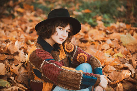 Young hipster woman in a fashionable knitted sweater while sitting on autumn foliage in the forest....