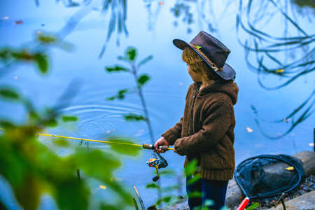 Little boy fishing in overalls from a dock on lake or pond. Child with a fishing rod standing by the...
