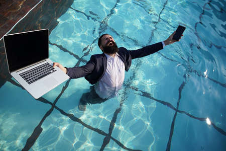 Crazy comic business. Excited business man in suit with laptop and mobile phone on swimming pool. Funny businessman relaxing with laptop.の写真素材