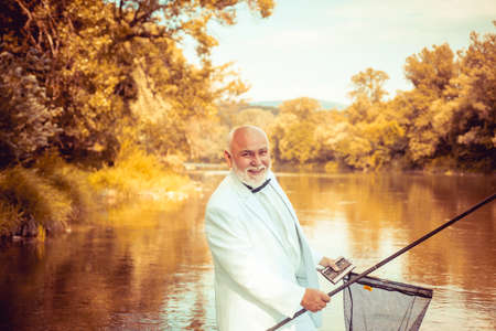 Fisherman in formal suit. Real happiness. Fishing background. Retirement fishery. Perfect weekend. Cheerful mature fisherman fishing in a river outdoors. Strategy. Fisherman fishing equipment.の写真素材