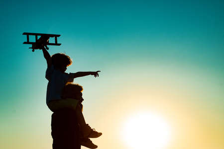 Sunset silhouette of Father carrying his son on shoulders. Child son pilot and dad daddy father with airplane.の写真素材