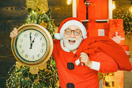 Five minutes to New year or Christmas midnight. Santa Claus. Cheerful Santa Claus holding alarm clock on fireplace and Christmas Tree background.の写真素材