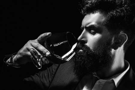 Cheerful bearded man is drinking expensive cognac. Old traditional whiskey drink - gentlemen beverage. Elegant and stylish man in classical wear holding glass with cognac in hand.の写真素材
