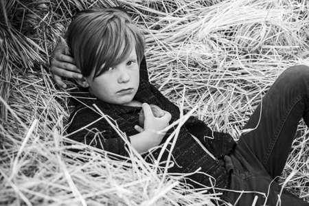 Kid boy holding gold leaf and lies on the hay. Fair-haired boy lies on hay background and eats an apple. Bye summer - hi autumn. Kid on an autumn holiday in the farm.の写真素材