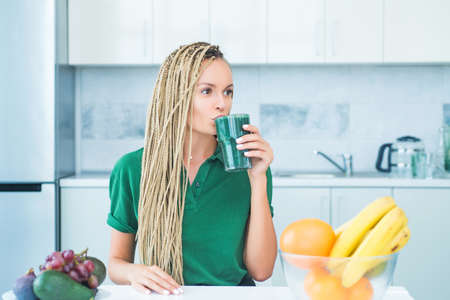 Young woman drinking green smoothie in the kitchen with fruits and vegetables. Healthy eating...