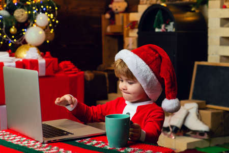 Smart toddler surfing internet. Little boy santa hat and costume. Boy child with laptop near christmas tree. Buy christmas gifts online. Christmas shopping concept. Gifts service. Santa little helperの写真素材
