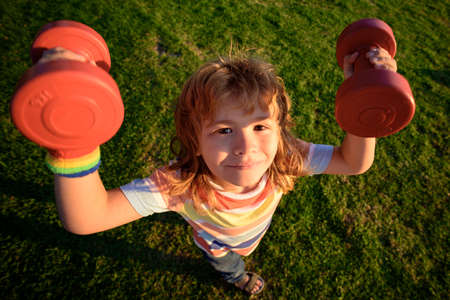 Funny child sport. Kid with dumbbells in park. Strong little boy. Fun child face. Children development and healthy strong exercise. Wide angle.の写真素材