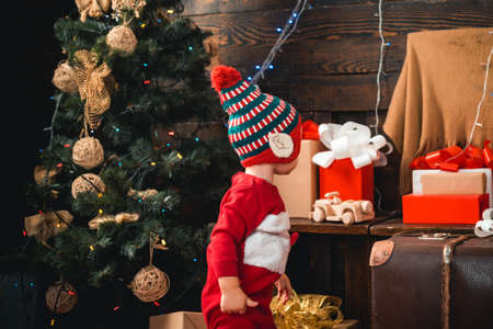 Babies. Happy small kids in santa hat with present have a christmas. Portrait kid with gift on wooden background. Winter kids. Portrait of happy child looking at decorative toy ball by Christmas tree.の写真素材